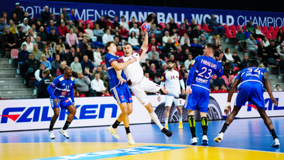 A handball player in a white uniform airborne while throwing a ball towards the goal past blue-jerseyed defenders, with blue Falken Tyre logos visible on the LED court barrier.