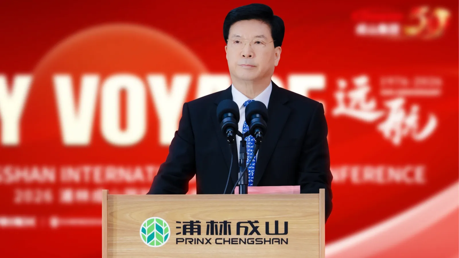 Che Hongzhi standing at a wooden podium speaking into microphones in front of a red 50th-anniversary backdrop.
