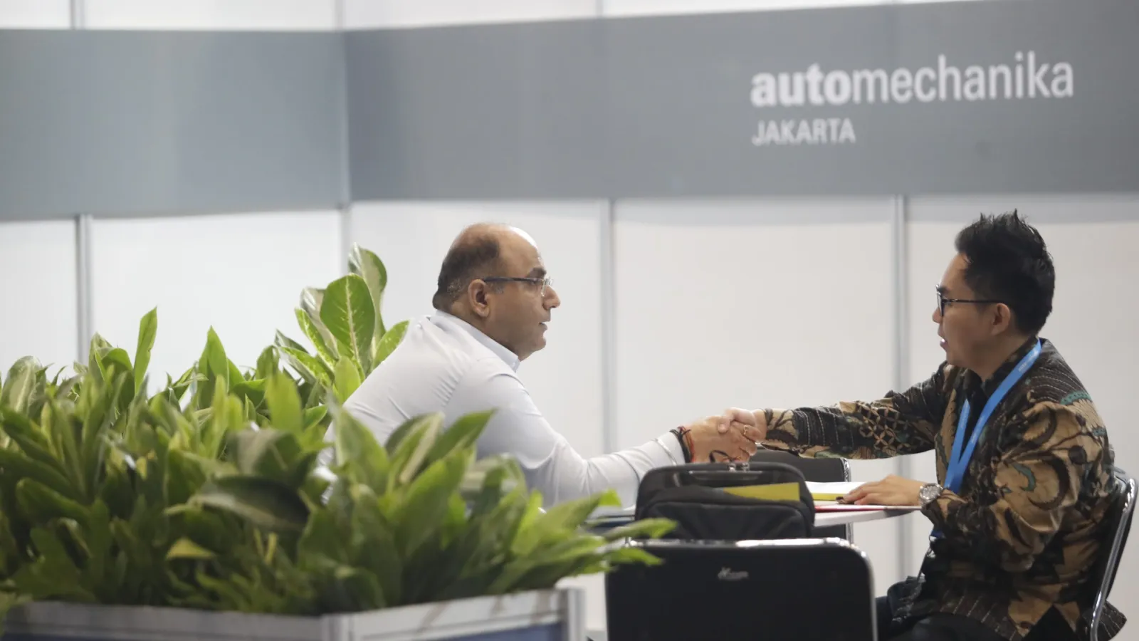 Two men in business attire shaking hands across a meeting table in front of potted green plants and a gray booth wall bearing the Automechanika Jakarta logo.